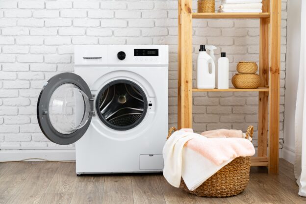 A modern front-loading dryer with the door open, placed against a white brick wall on a wooden floor. Beside it is a wooden shelf stocked with laundry detergents, towels, and woven baskets. A wicker laundry basket filled with folded towels is in the foreground.