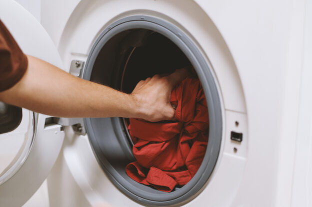 A close-up of a person’s hand placing a red shirt into the drum of a front-loading dryer machine. The open door reveals the machine’s gray rubber seal and white exterior, indicating the start of a laundry cycle. Dryer duct maintenance.